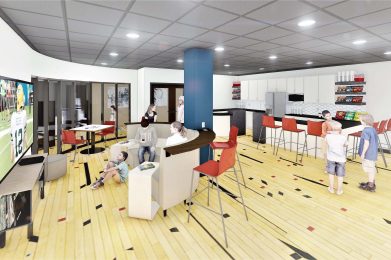 room with seating, a TV, and kitchenette, showing people engaged in leisure activities.